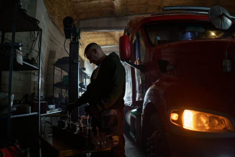 A mechanic focuses on his work beside a large red truck in a dimly lit workshop.